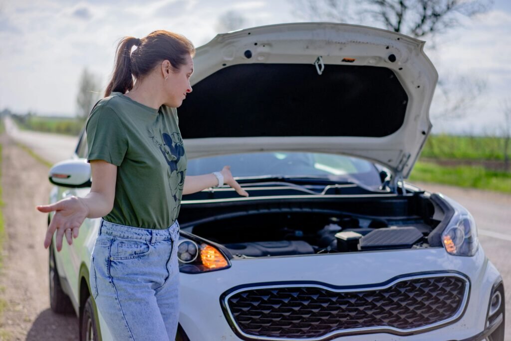 pexels-photo-12956065-12956065 A woman looks puzzled by her broken car with an open hood on a rural road.
