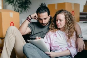 pexels-photo-4246012-4246012 Pensive male and female in casual clothes sitting together among boxes and writing notes in notebook while leaning on bed